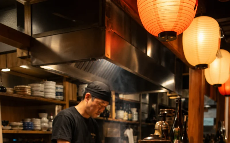 A wide-angle, photorealistic shot inside a warm-lit Tokyo izakaya , wooden counter seats, glowing paper lanterns, ceramic sake cups, and small plates of grilled skewers , no phone numbers or brand signage visible. Alt: best izakaya in Tokyo interior atmosphere with sake and yakitori.