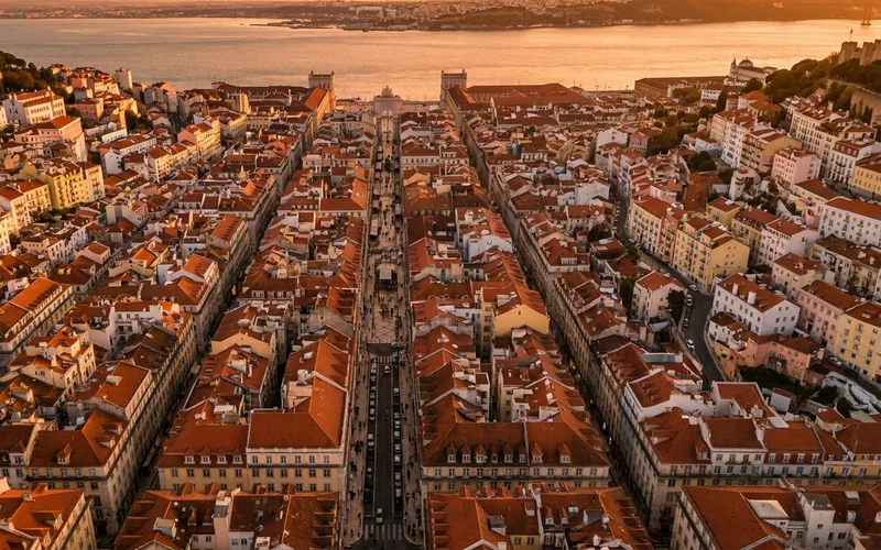 A photorealistic 16:9 aerial view of central Lisbon at golden hour, showing the terracotta rooftops, the Tagus River shimmering in the background, and the grid-like streets of Baixa meeting the hillside neighborhoods. Alt: Best neighborhoods to stay in Lisbon aerial overview at sunset.